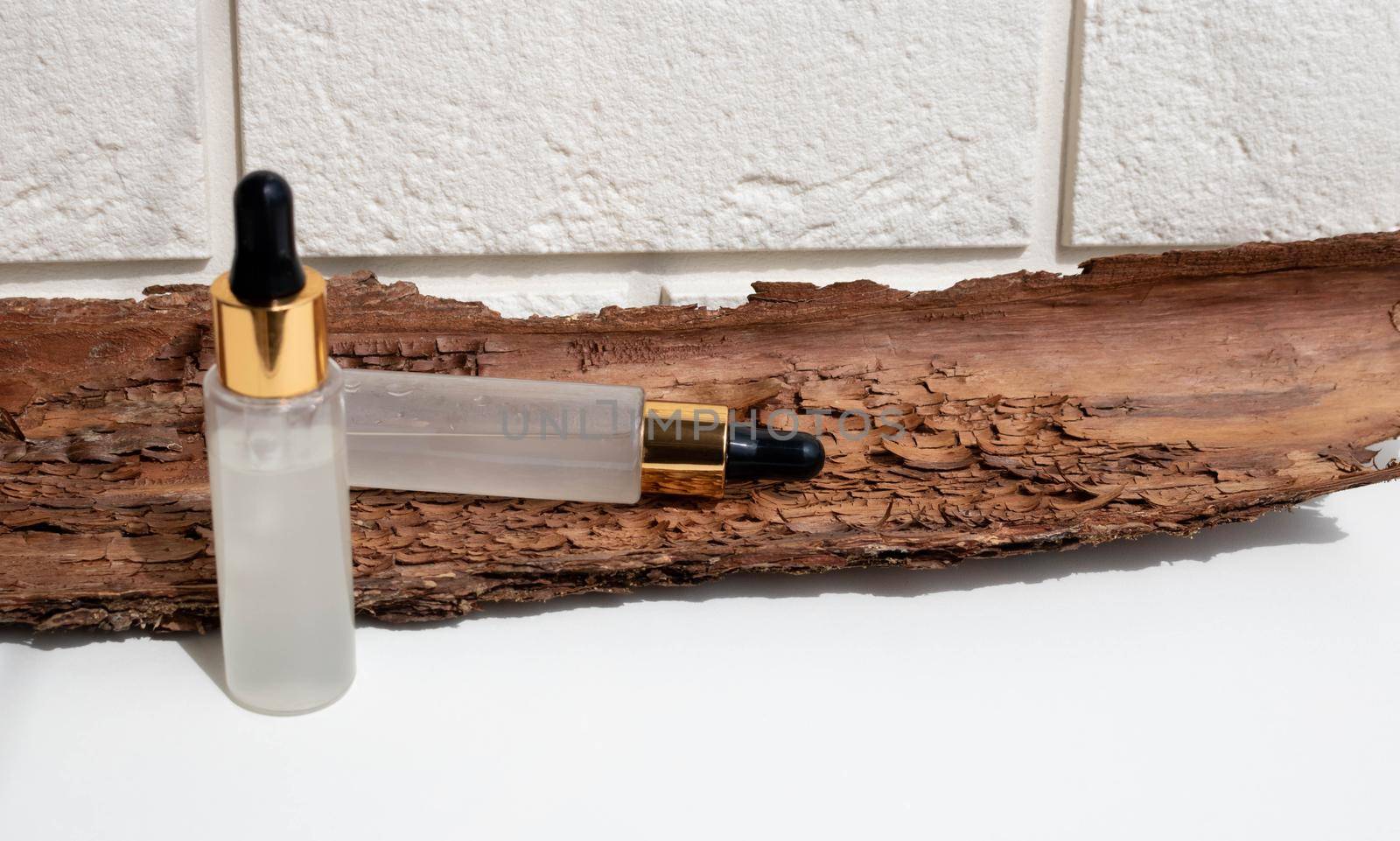 A bottle of serum with a pipette on a background of white brick and brown bark. A close-up of a frosted glass container for a cosmetic skin care product