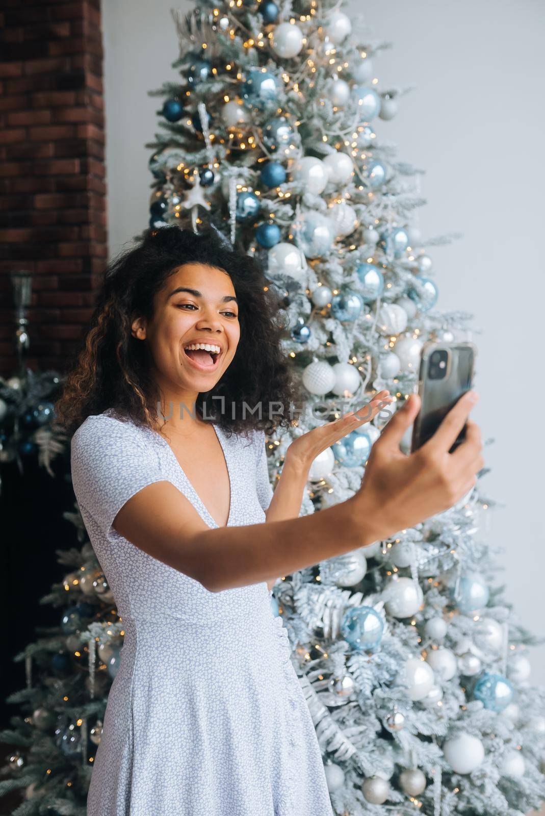 Beautiful Young Smiling Woman near Christmas Tree at the Background Making Video Message or Selfie Concept of Holidays and New Year in Christmas decoration room.