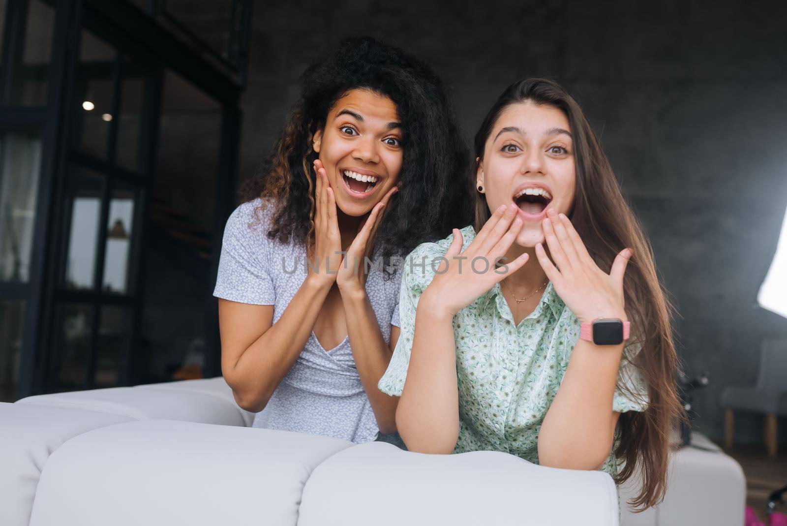 Two girls sit on the back of the sofa, look at the cameras and smile at home