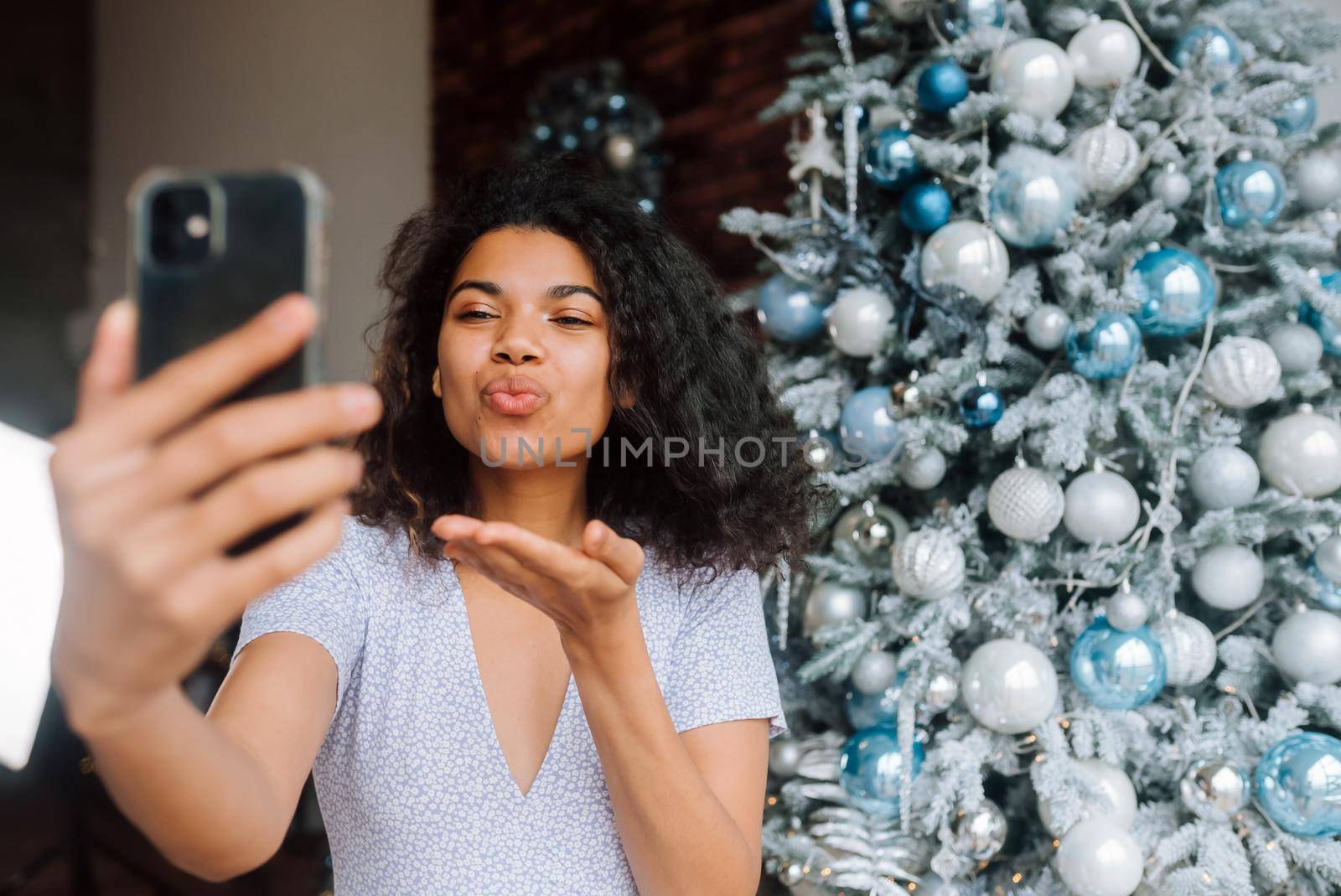 Beautiful Young Smiling Woman near Christmas Tree at the Background Making Video Message or Selfie Concept of Holidays and New Year in Christmas decoration room.