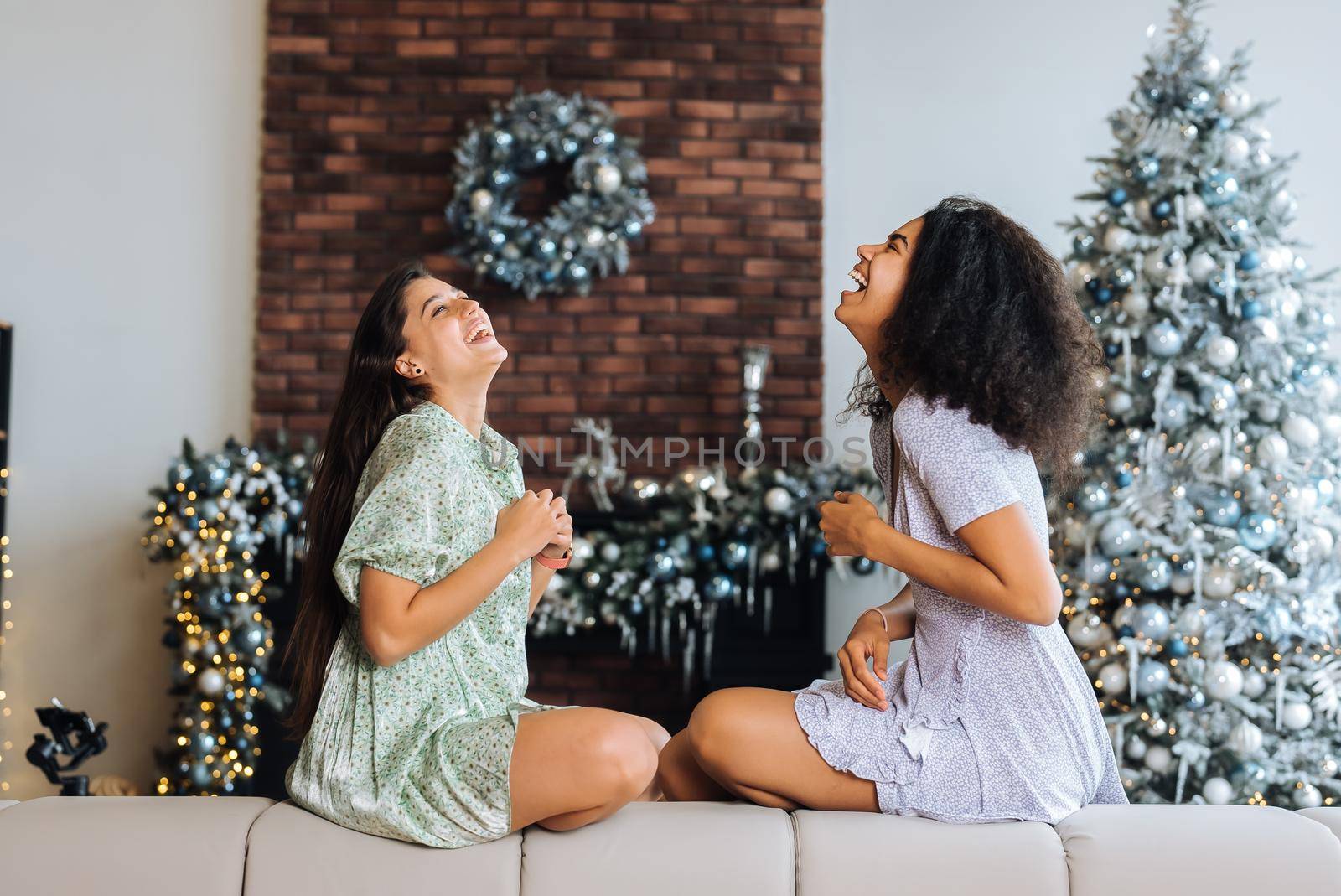 Two girls sit on the back of the sofa, look at the cameras and smile at home