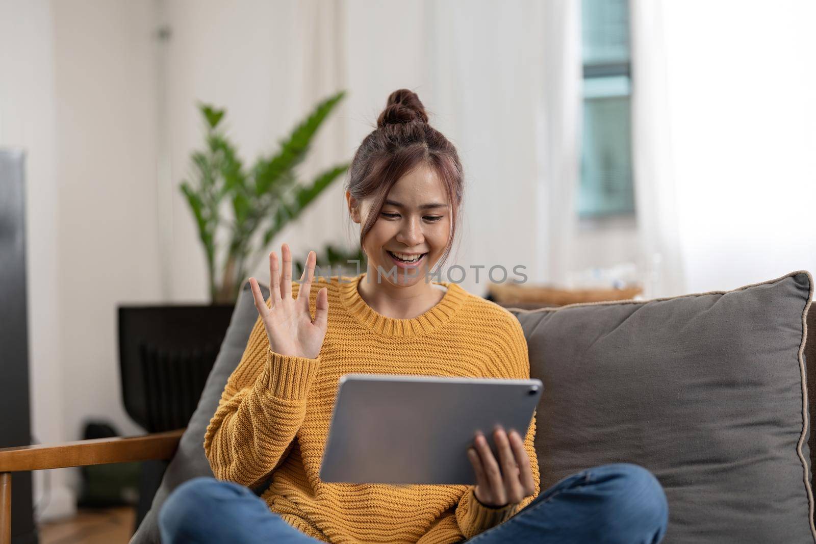 Happy smiling asian young woman in yellow sweater with tablet pc computer having video call and waving hand at home.
