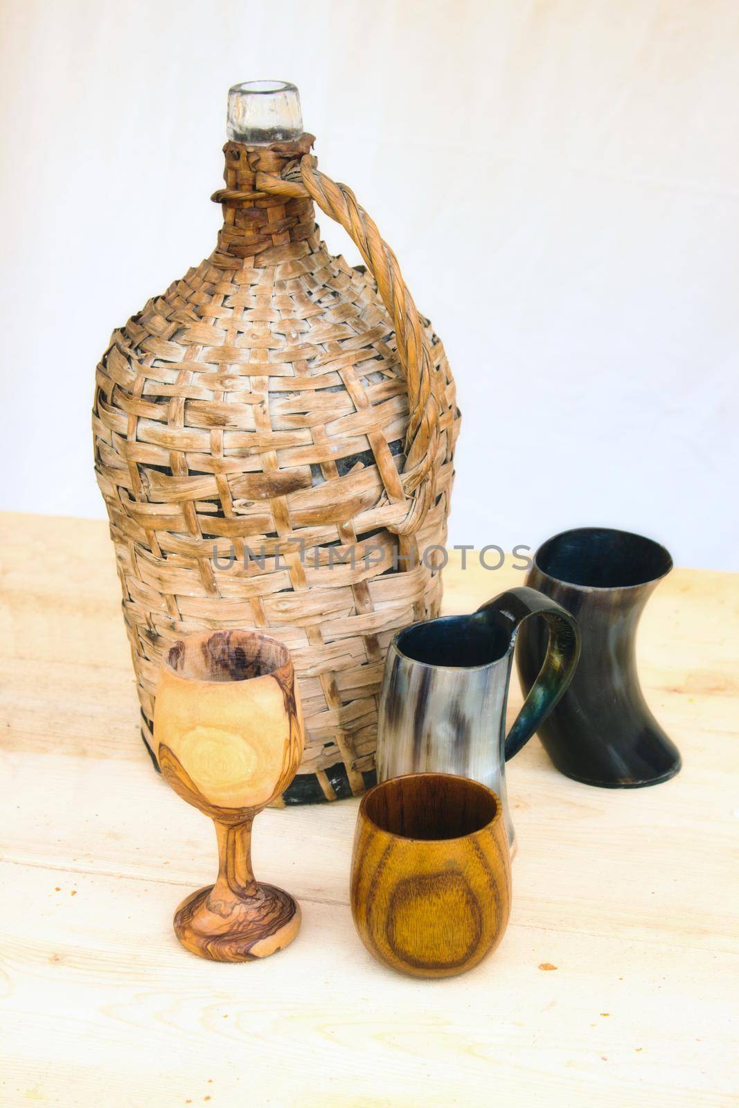 A traditional glass bottle encased in wicker with wooden cups on a table