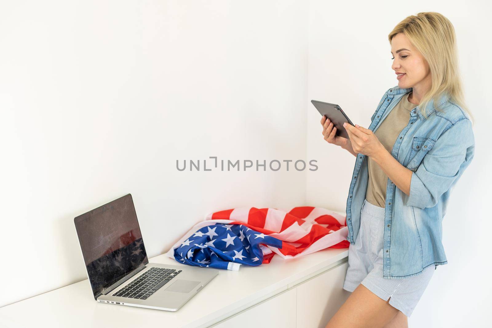 Happy young female student hold flag of USA, studying with laptop with blank screen in living room interior. International education at home, lesson remote, website due covid-19 quarantine by Andelov13