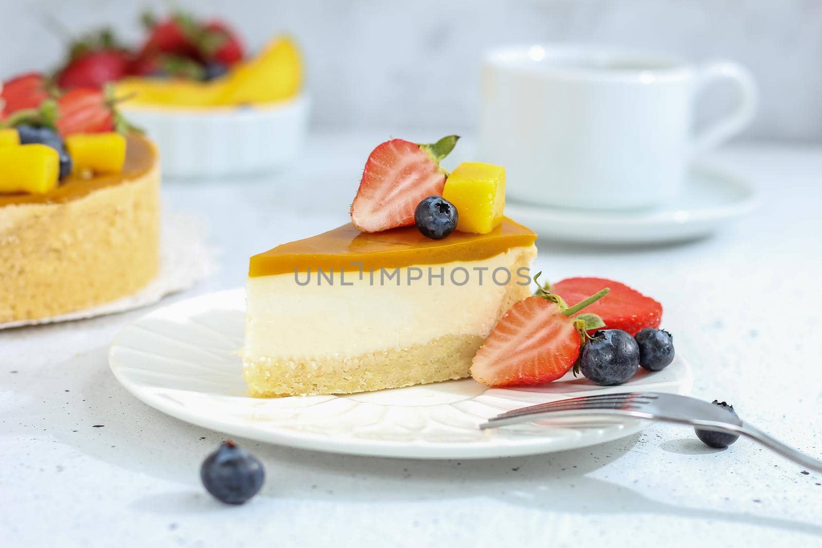 A piece of cheesecake with mango on a plate with a tea fork decorated with berries and flowers on a gray background. Healthy food.