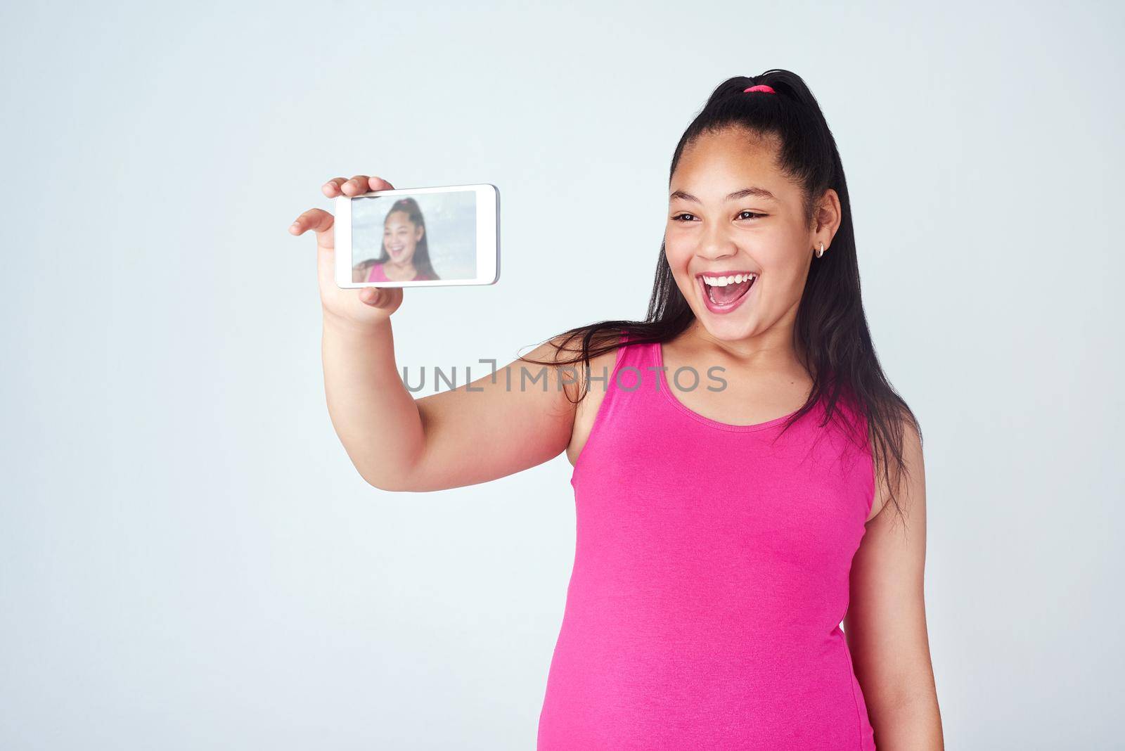 Studio shot of a cute young girl taking a selfie with a mobile phone against a gray background.
