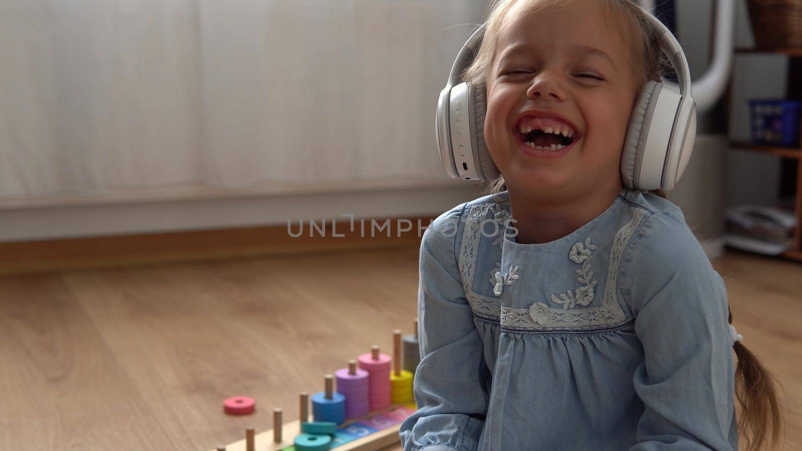 Child Listening Music In Big White Headphones. Happy Little Preschool Toothless Girl 5 Years Old Looking At Camera In Leaving Room Inside. Kid Smiles Laughs Shows Tongue At Home. Childhood, Education.