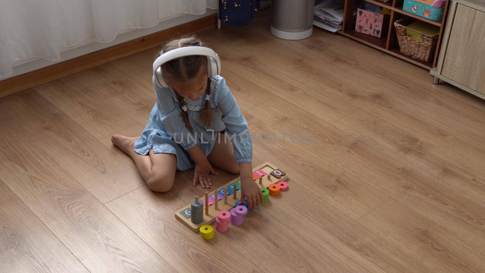 Happy Little Preschool Toothless Girl Playing With Colored Wooden Toy. Kids Learn To Count By Playing Teaches Numbers At Home. Child Listening To Music In Big White Headphones. Childhood, Education.