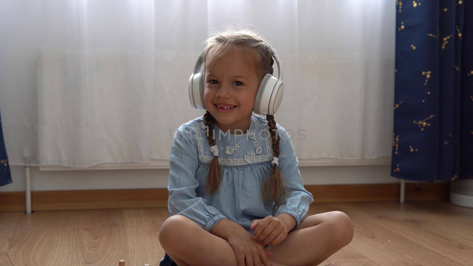 Child Listening Music In Big White Headphones. Happy Little Preschool Toothless Girl 5 Years Old Looking At Camera In Leaving Room Inside. Kid Smiles Laughs Shows Tongue At Home. Childhood, Education.