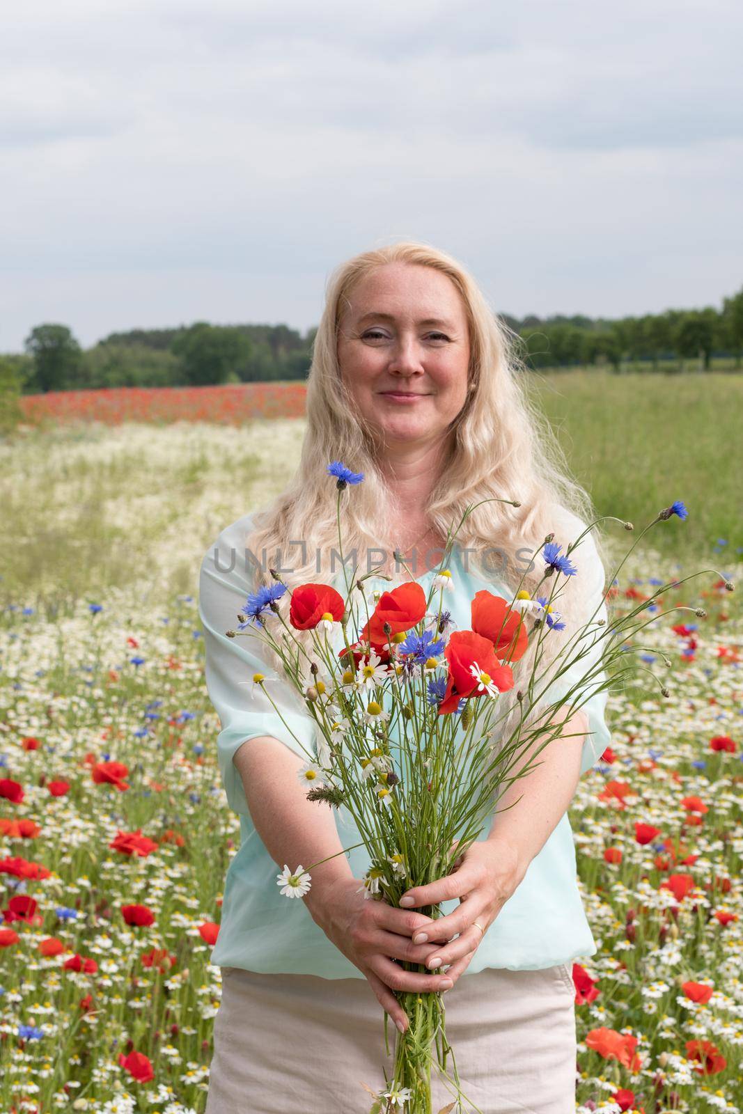 a beautiful middle-aged blonde woman stands among a flowering field of poppy, daisies, Cornflowers, and holds a bouquet of wild flowers and laughs. High quality photo