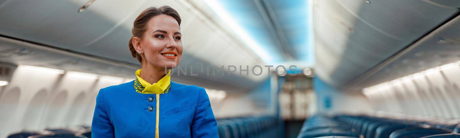 Cheerful woman stewardess in airline air hostess uniform standing in aisle of airplane passenger salon