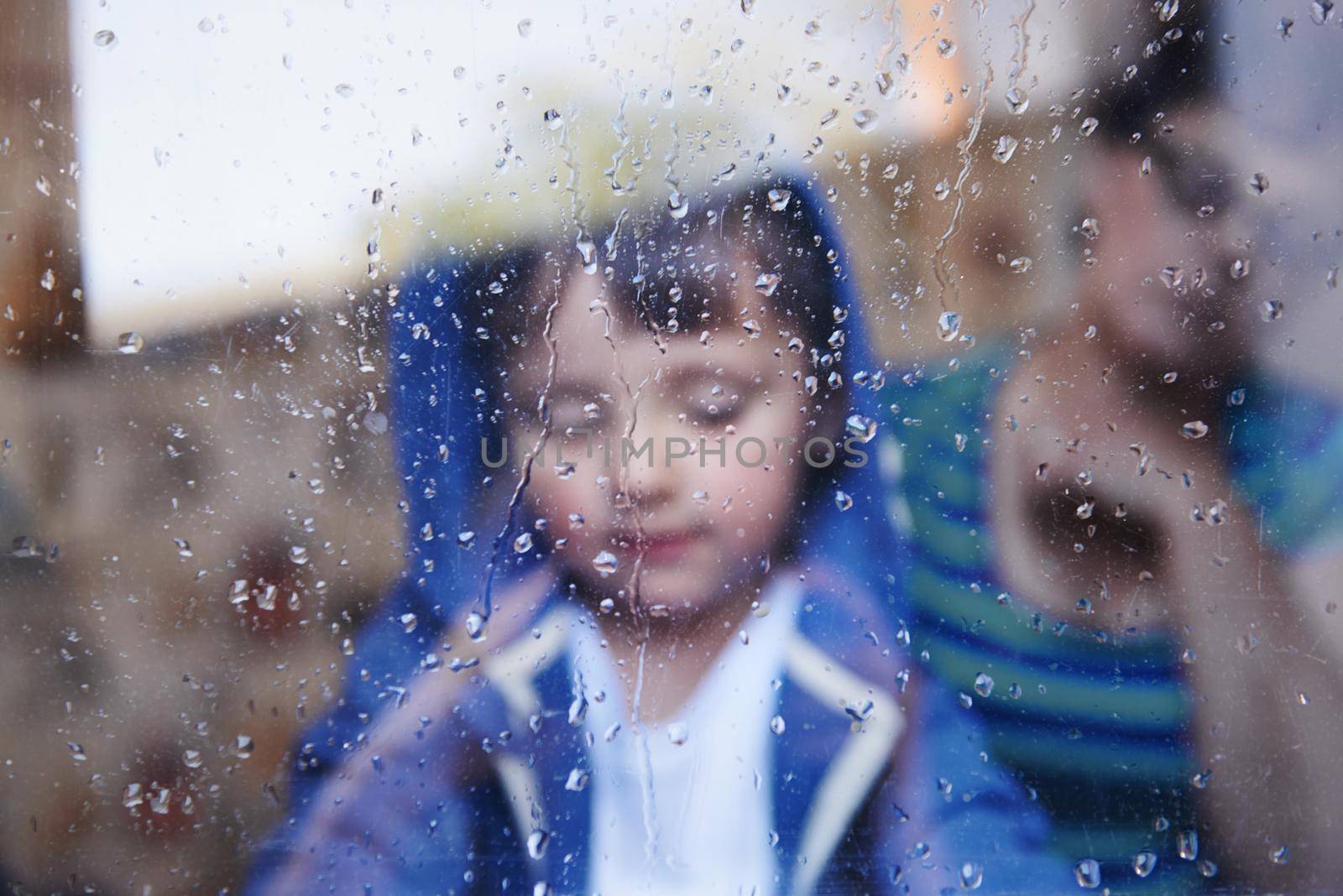 Rainy days with nothing to do.... A little boy looking downwards behind a rain-streaked window while his mother sits in the background. by YuriArcurs