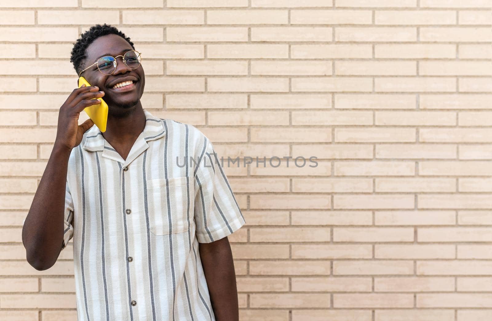 Smiling, happy African American man using mobile phone to talk. Black male on phone call in the street. Brick wall background. Copy space. Lifestyle and technology concept.