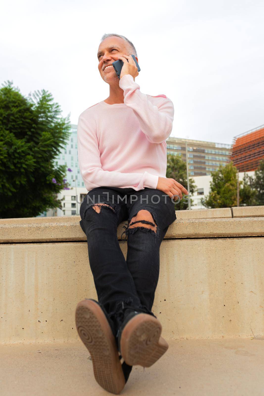 Smiling caucasian man talking on mobile phone sitting on stairs in the city. Vertical image. by Hoverstock