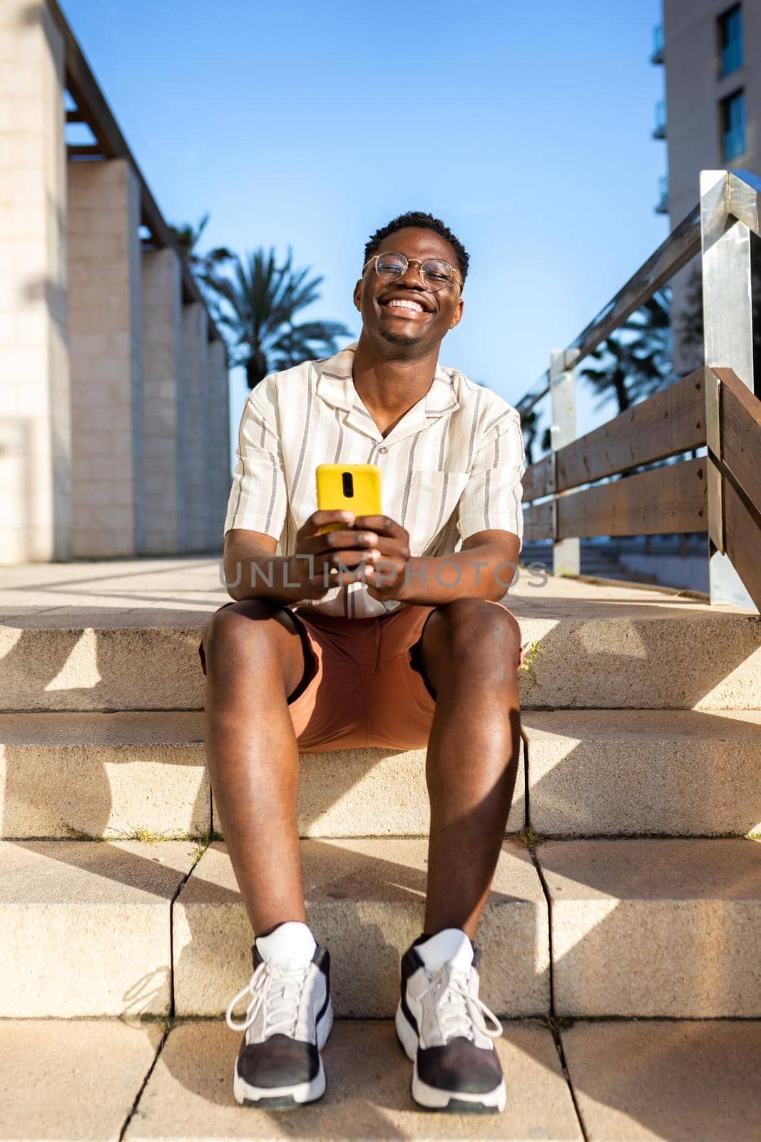 Happy, young African American man using mobile phone outdoors sitting on stairs. Vertical image. by Hoverstock