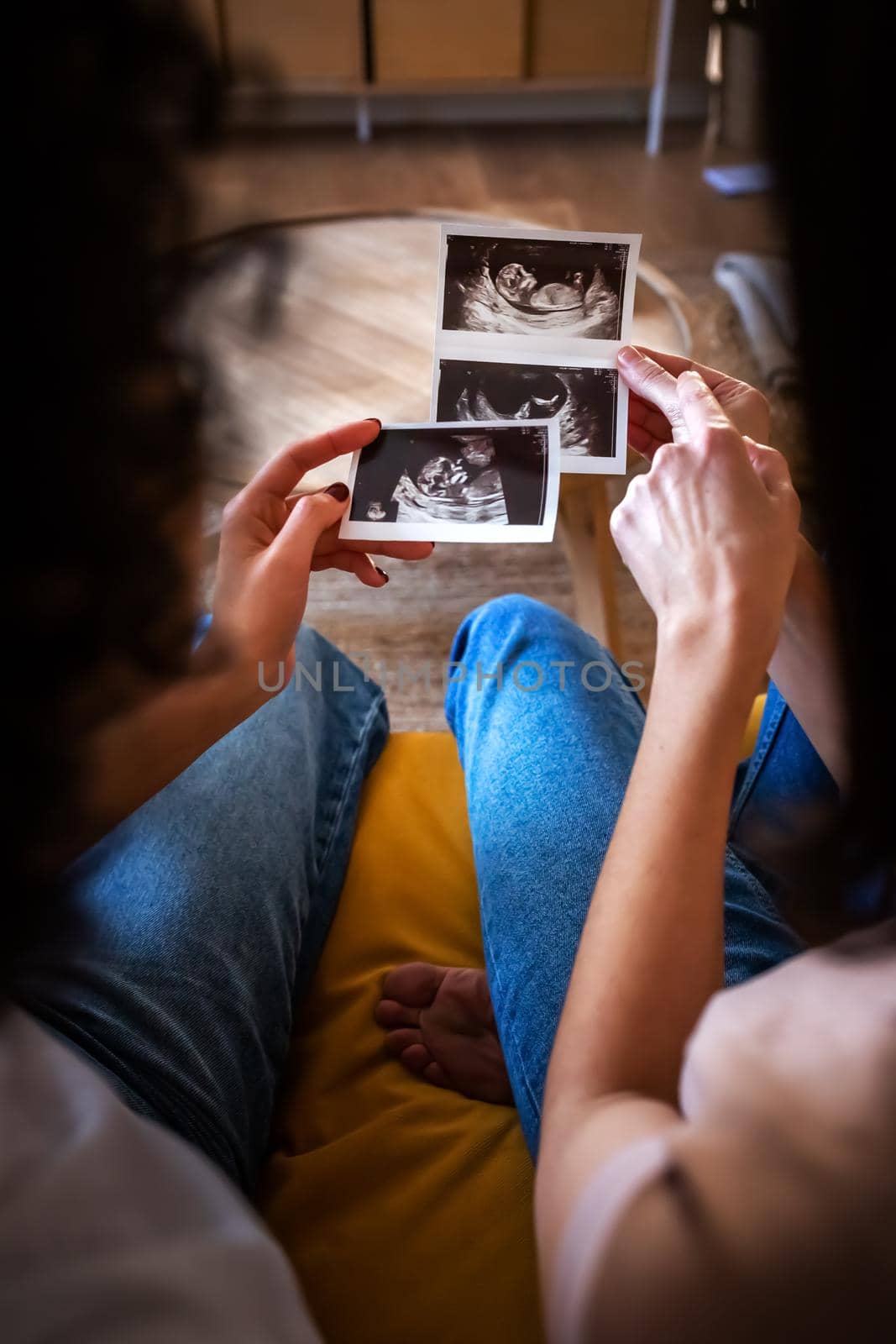 Top view of multiracial lesbian pregnant couple looking at each other in love while holding baby ultrasounds. Vertical image. LGTBQ love and family concepts.