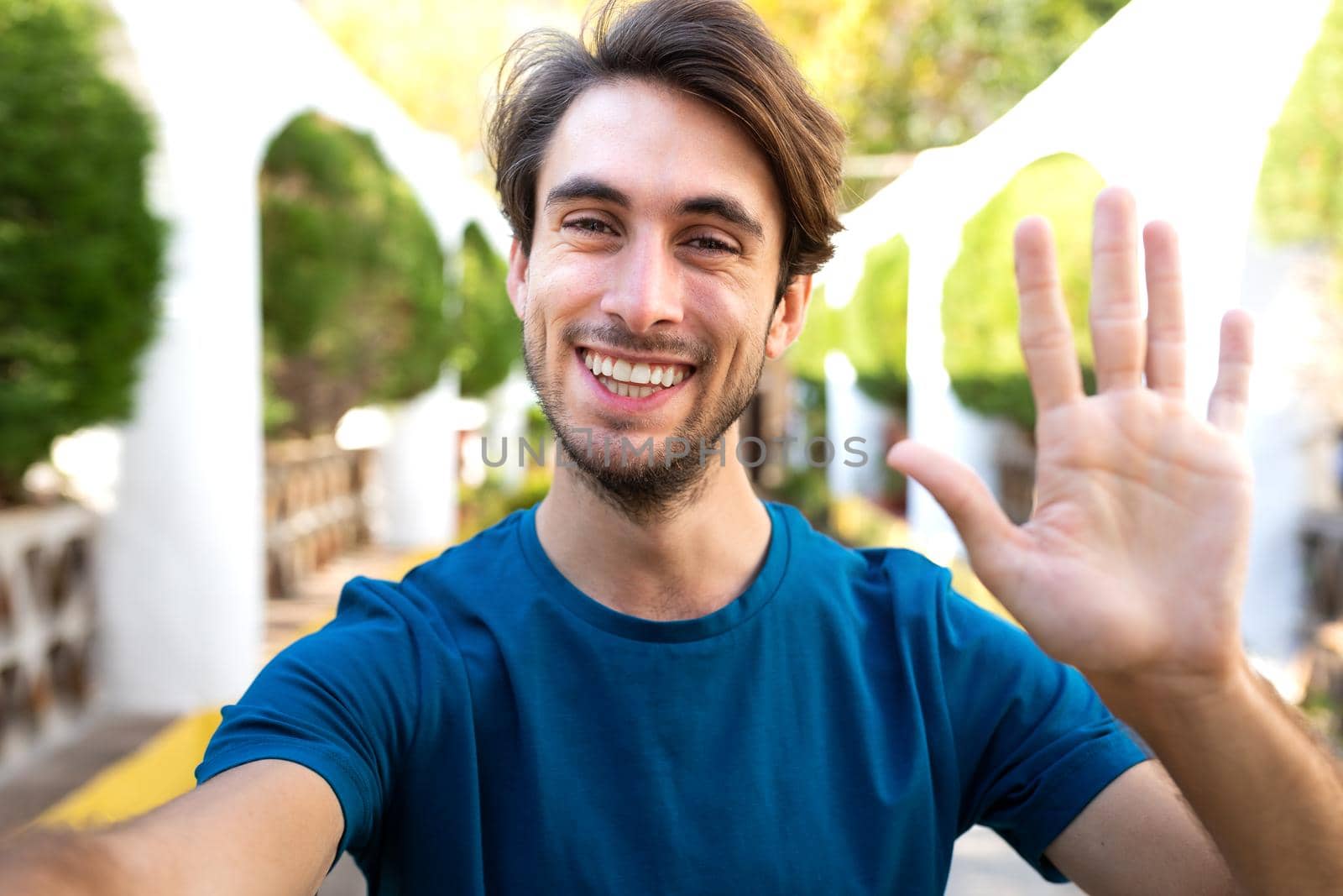 Young caucasian man wave hello on a video call. Male take selfie in city park. by Hoverstock