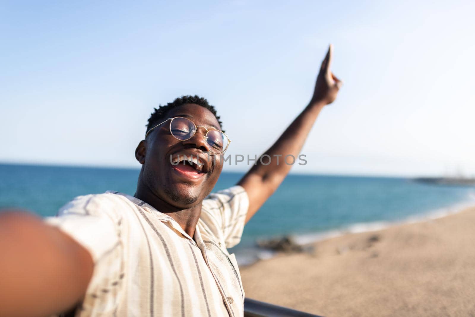Ecstatic African American man taking selfie at the beach during vacations. Smiling, happy, Black male with glasses looking at camera waving hello. Vacation concept.