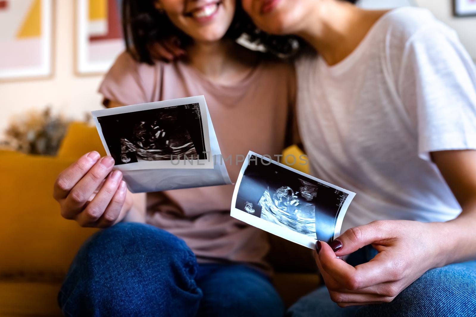 Close up of unrecognisable happy multiracial pregnant lesbian couple looking baby ultrasounds at home. LGTBQ love and family concept.