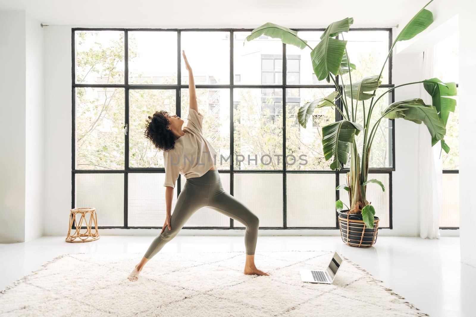 Young multiracial woman practicing yoga at home with online lesson using laptop. Reverse warrior pose. Copy space. by Hoverstock
