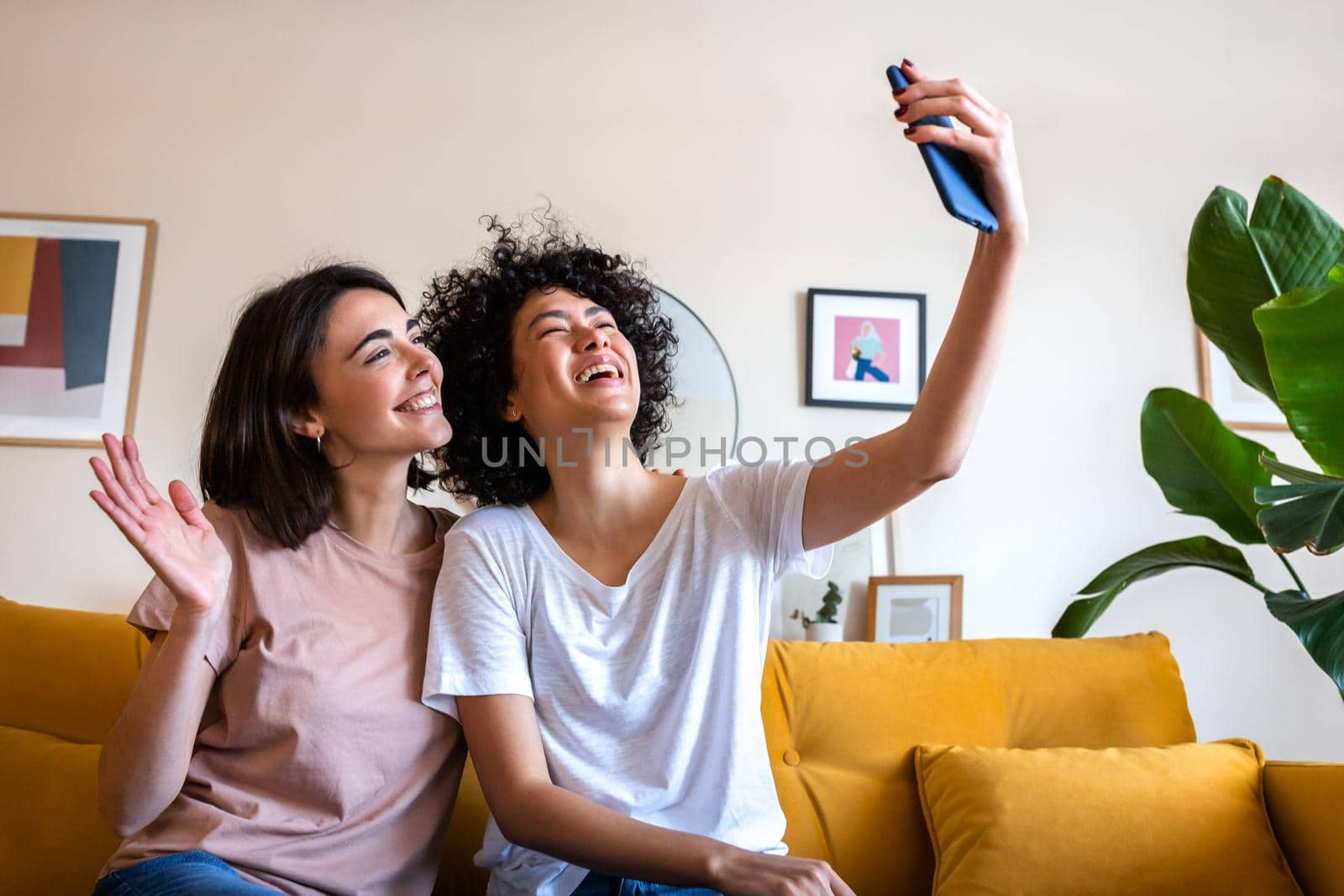 Multiracial gay female couple sitting on the couch at home waving hand during video call using mobile phone. Lgbt and technology concept.