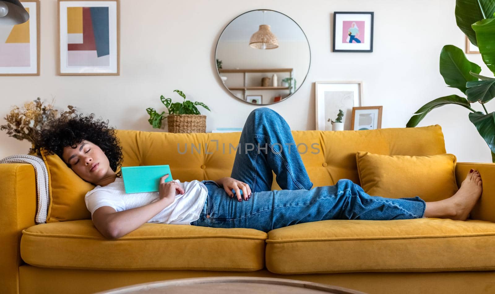 Tired African American woman sleeping on yellow couch after reading a book Female relaxing at home. by Hoverstock