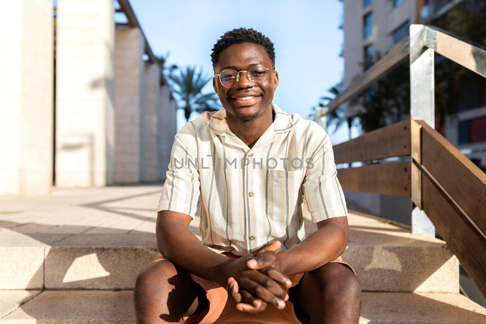 Happy African American young man sitting on stairs outdoors looking at camera. by Hoverstock