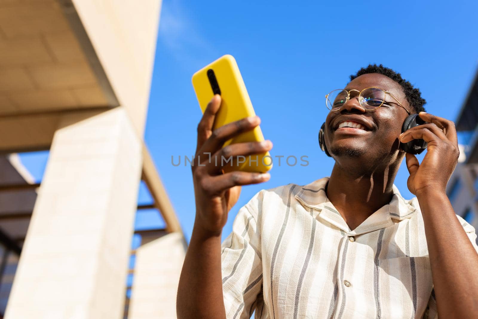 Black young man relaxing outdoors listening to music using phone and headphones. Copy space. by Hoverstock