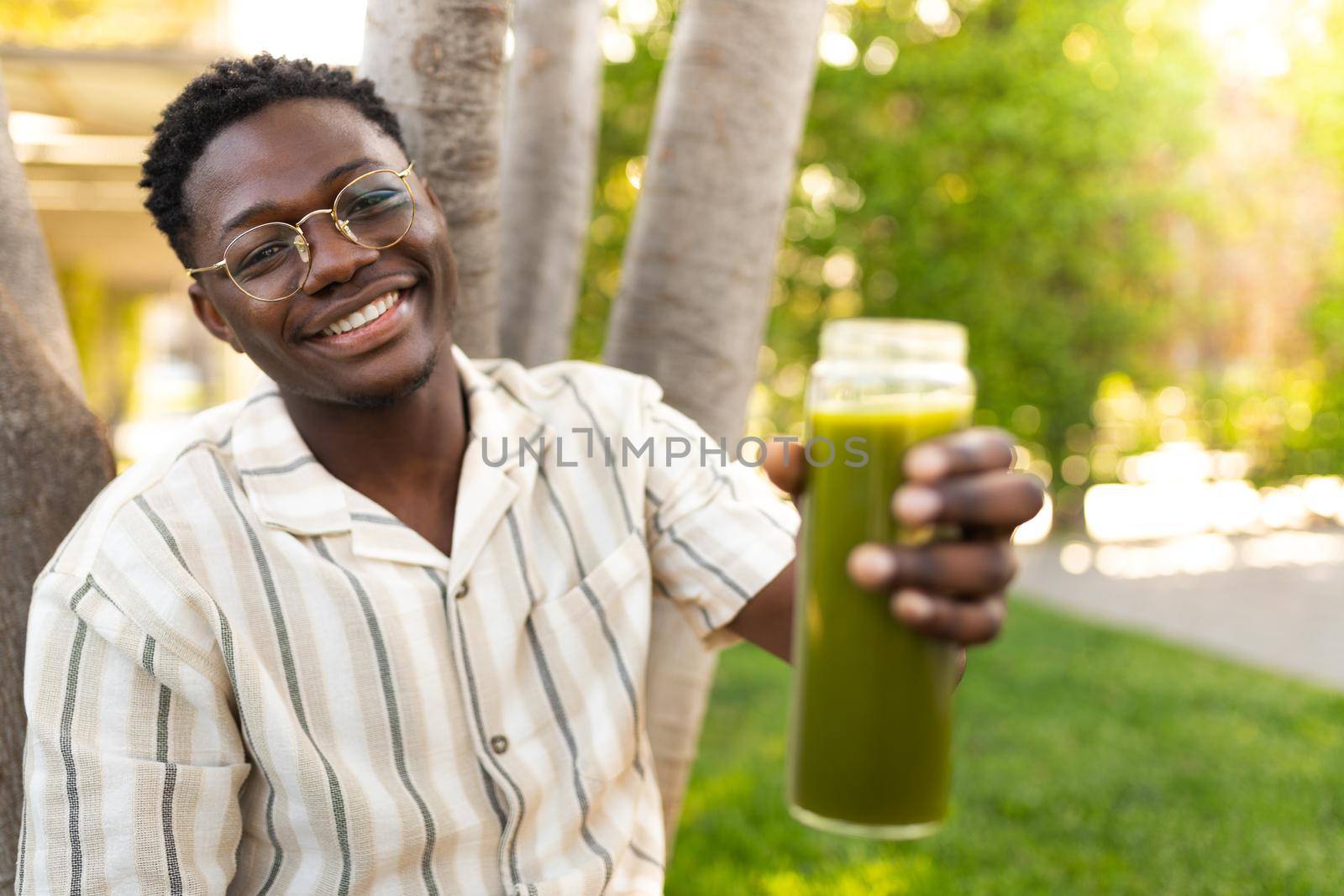 Young black man showing glass bottle of green juice to camera. Focus on man face. by Hoverstock