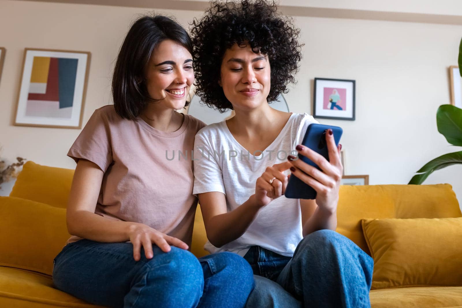 Young multiracial lesbian couple sitting on the couch looking at mobile phone together. Gay couple checking social media. Lgbt people and technology concept.