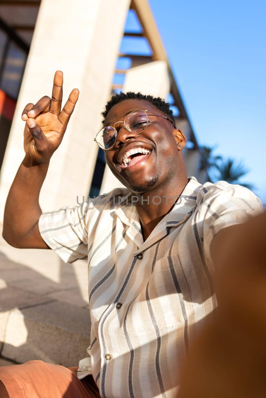 African american man with glasses taking selfie outdoors looking at camera making peace sign. Vertical image. Lifestyle concept.