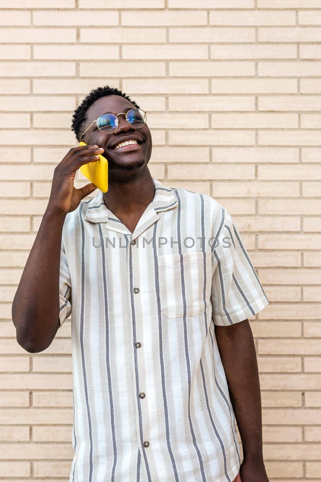 Vertical portrait of smiling, happy black man using mobile phone. African american male on phone call in the street. Brick wall background. Lifestyle and technology concept.
