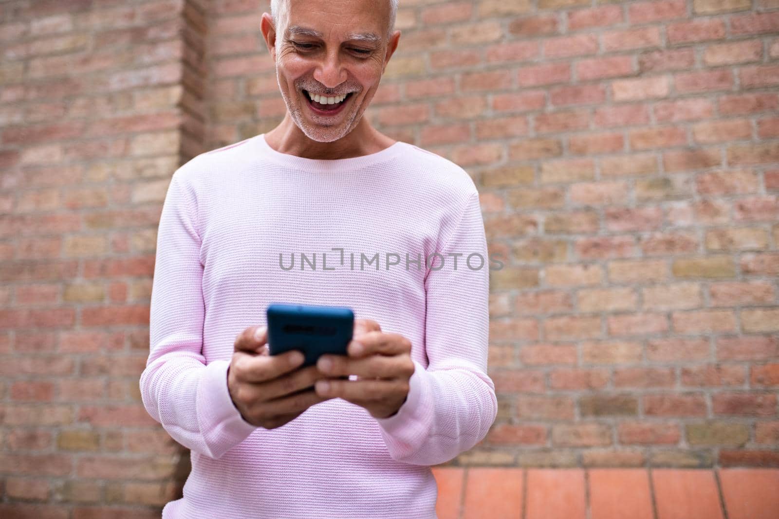 Smiling caucasian adult man looking at cellphone. Browsing through social media. Orange brick wall background. Copy space. Lifestyle and technology concept.