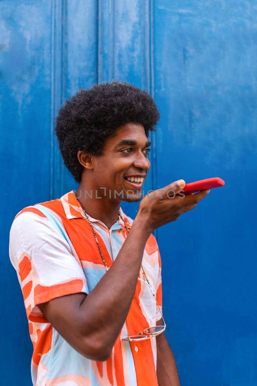 Happy black man with afro haistyle recording voice note using mobile phone in the street. African American man using phone to send voice message. Vertical image. Technology concept.