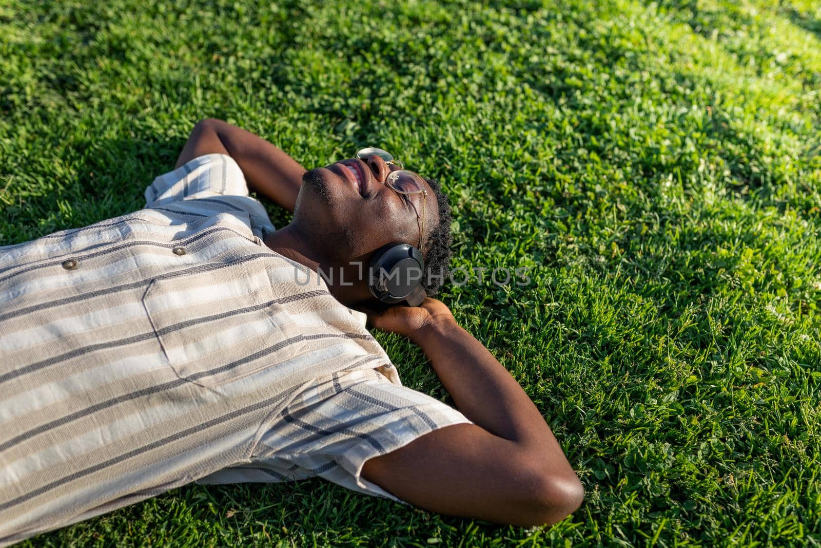 Black man relaxing outdoors. Happy black male lying on grass listening to music with headphones. Copy space. by Hoverstock