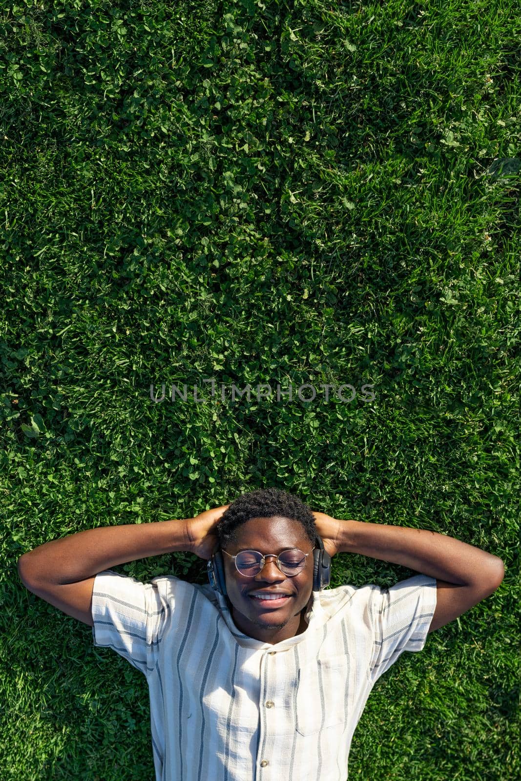 Top view of black man relaxing. Happy black male lying on grass listen to music with headphones. Copy space Vertical by Hoverstock