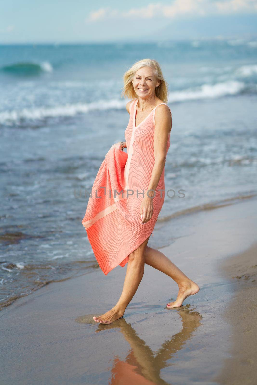 Beautiful senior woman walking along the shore of a tropical beach, wearing a nice orange dress. Elderly female enjoying her retirement at a seaside retreat.