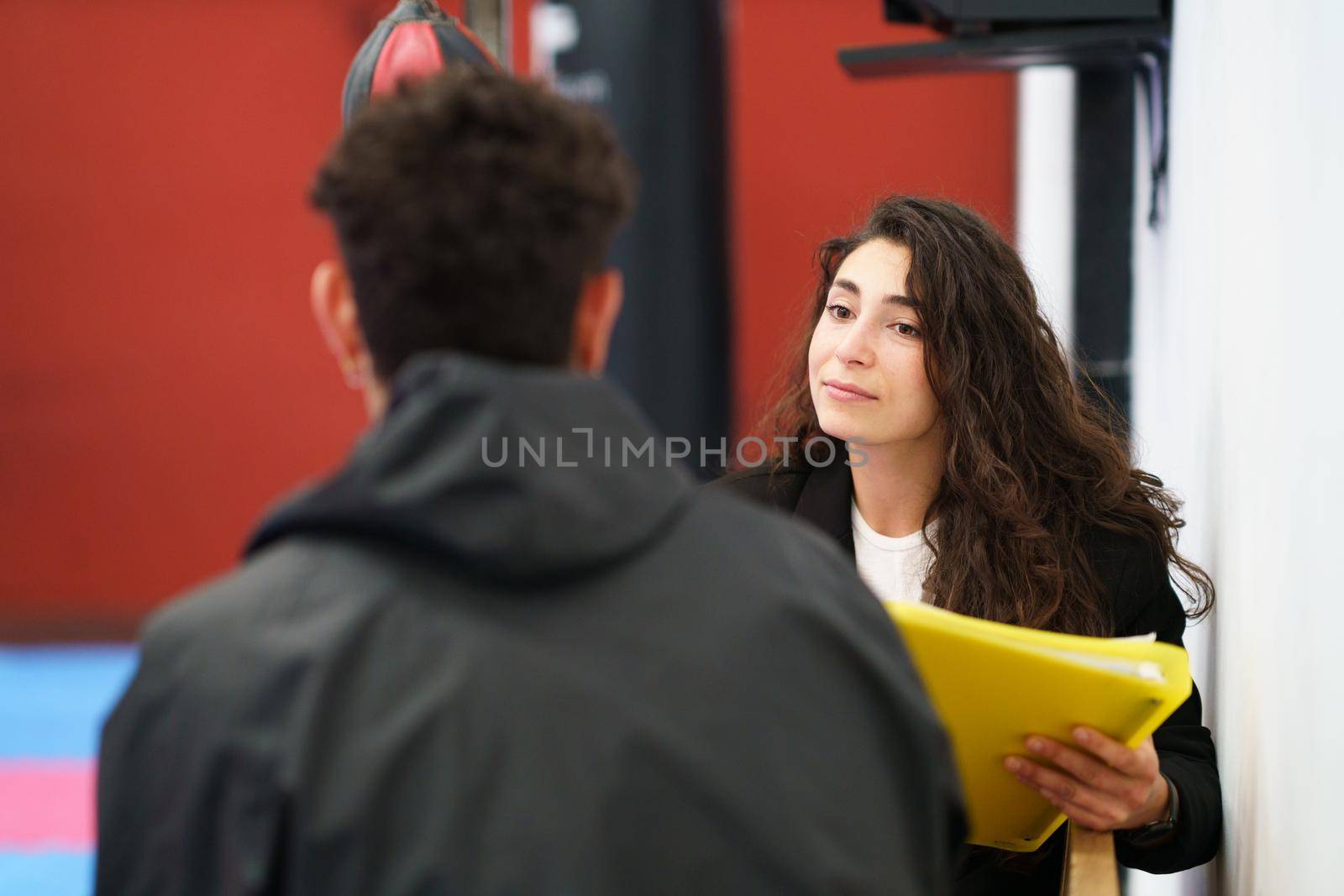 Young woman with yellow folder listening to sportsman while sitting on bench during motivation therapy session in modern gym