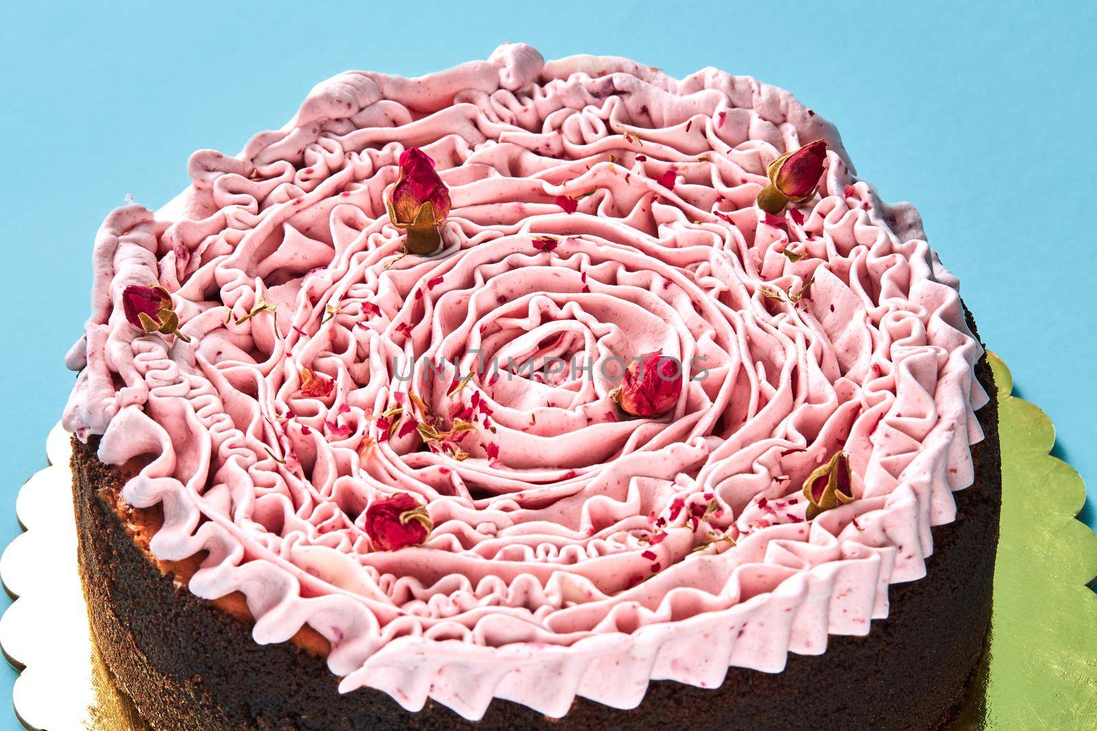 Close-up shot of a pink cream cake decorated with a small dried rosebuds, standing on a gold stand against a blue studio background. Pastries, delicious holiday bake.