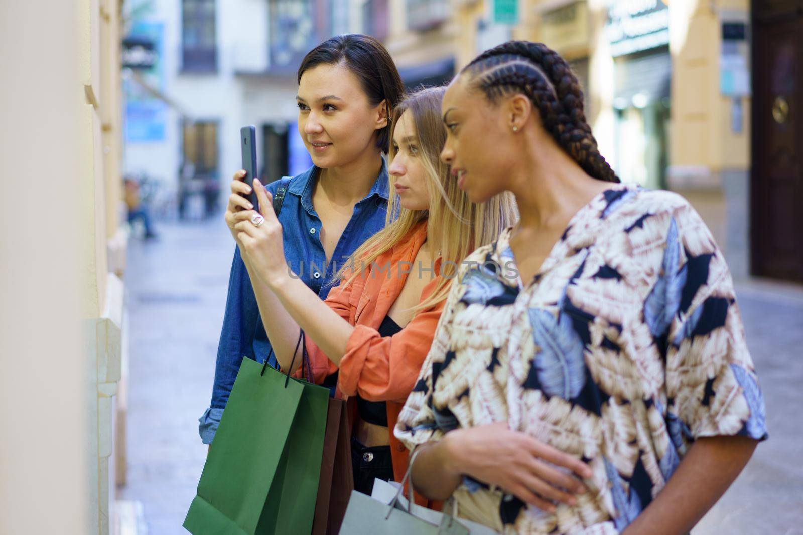 Side view of multiethnic female friends with shopping bags taking picture of showcase on smartphone while standing near store in city