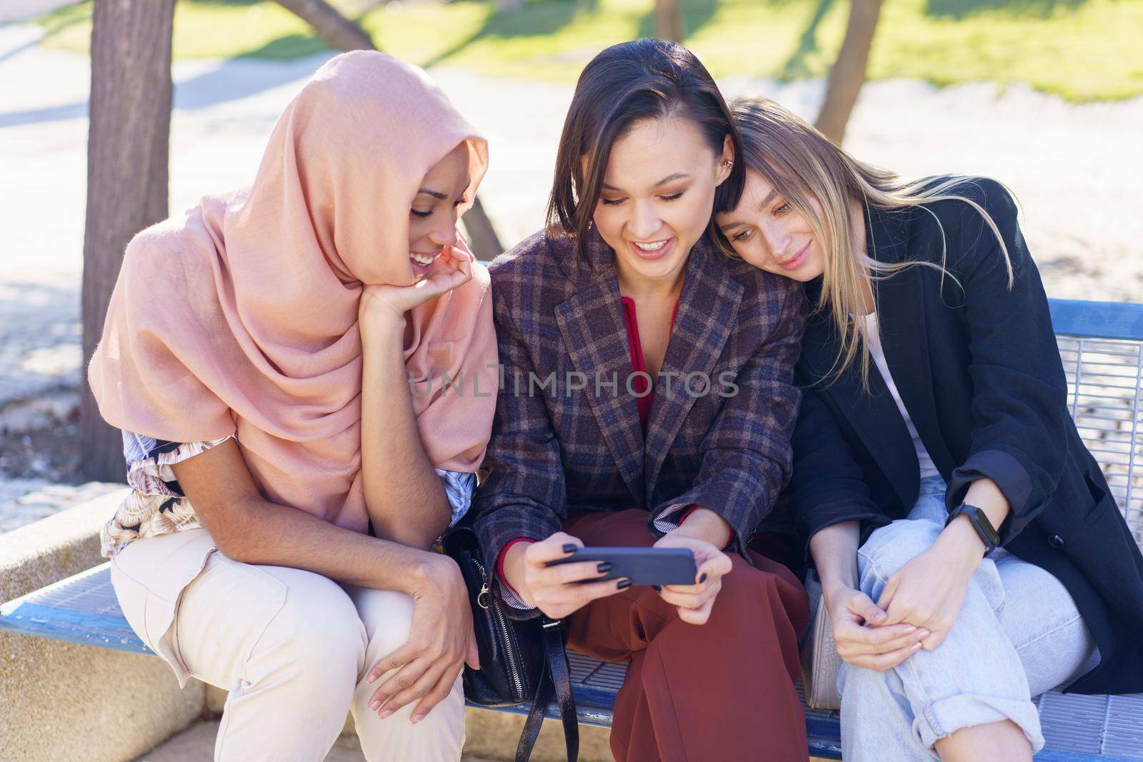 Smiling young diverse ladies watching video on smartphone in park by javiindy