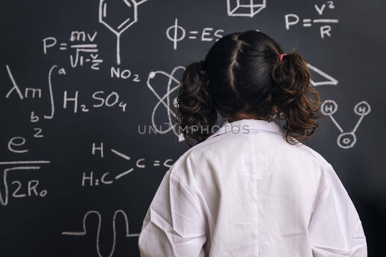 rear view of a little girl science student in lab coat on school blackboard background with hand drawings science formula pattern, back to school and successful female career concept