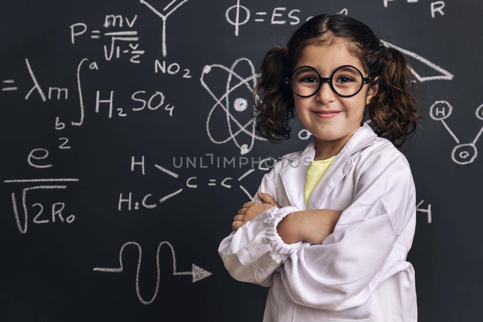 cheerful little girl science student in lab coat on school blackboard background with hand drawings science formula pattern, back to school and successful female career concept