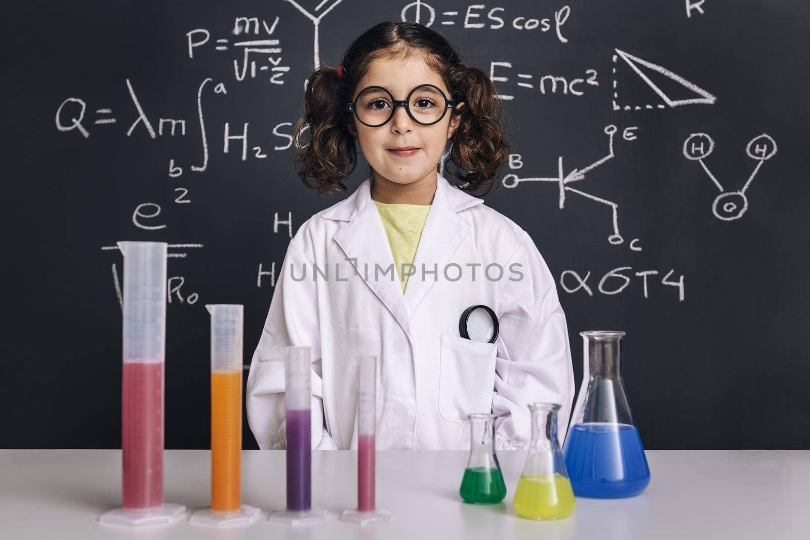 little scientist girl with glasses in lab coat with chemical flasks, on school blackboard background with hand drawn science formulas, back to school and successful female career concept