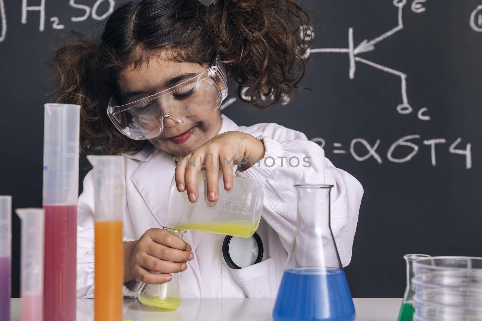 funny scientist child with goggles and gloves in lab coat mixing chemical liquids in flasks, blackboard background with science formulas, explosion in the laboratory, back to school concept