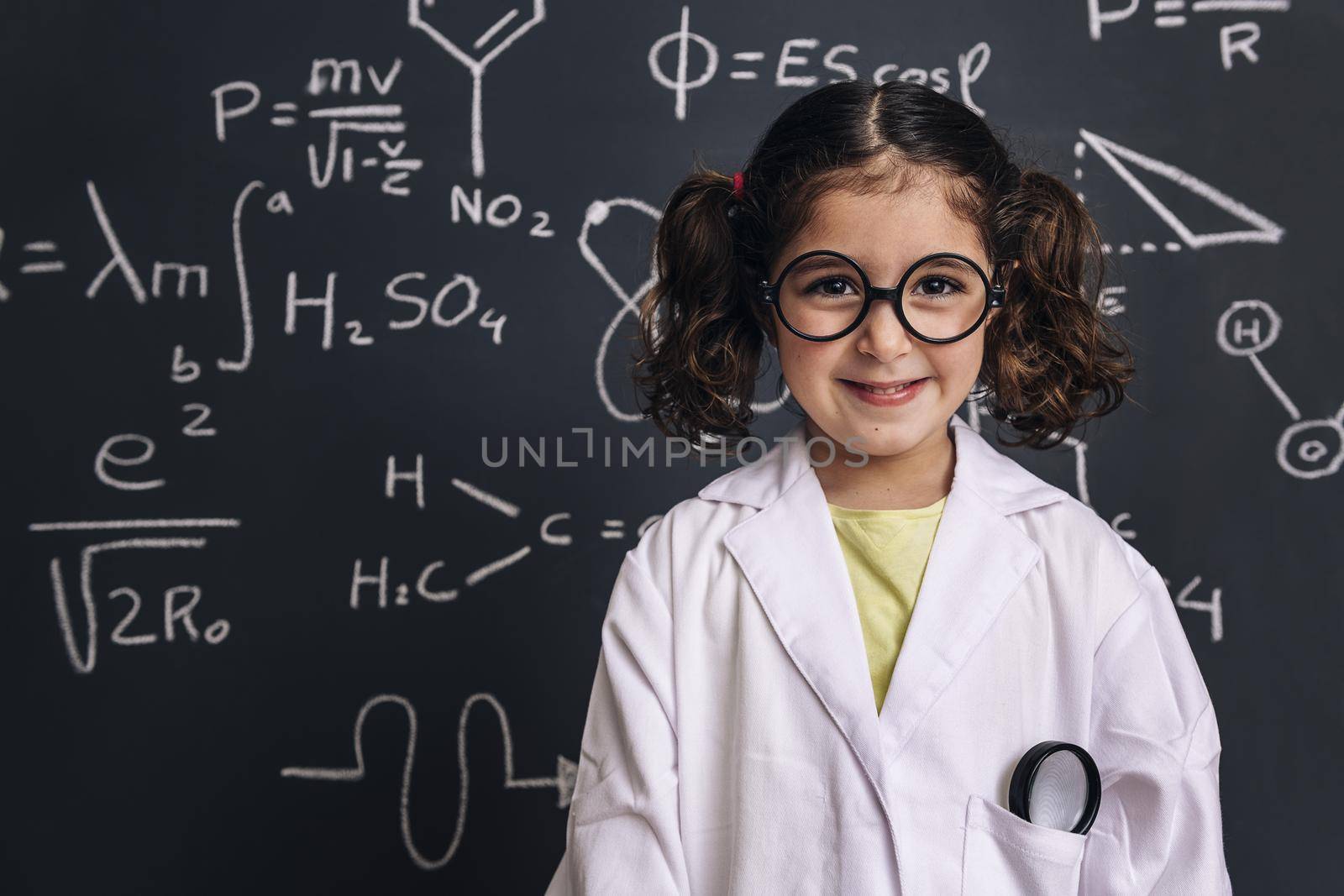 cheerful little girl science student with glasses in lab coat smiles on school blackboard background with hand drawings science formula pattern, back to school and successful female career concept