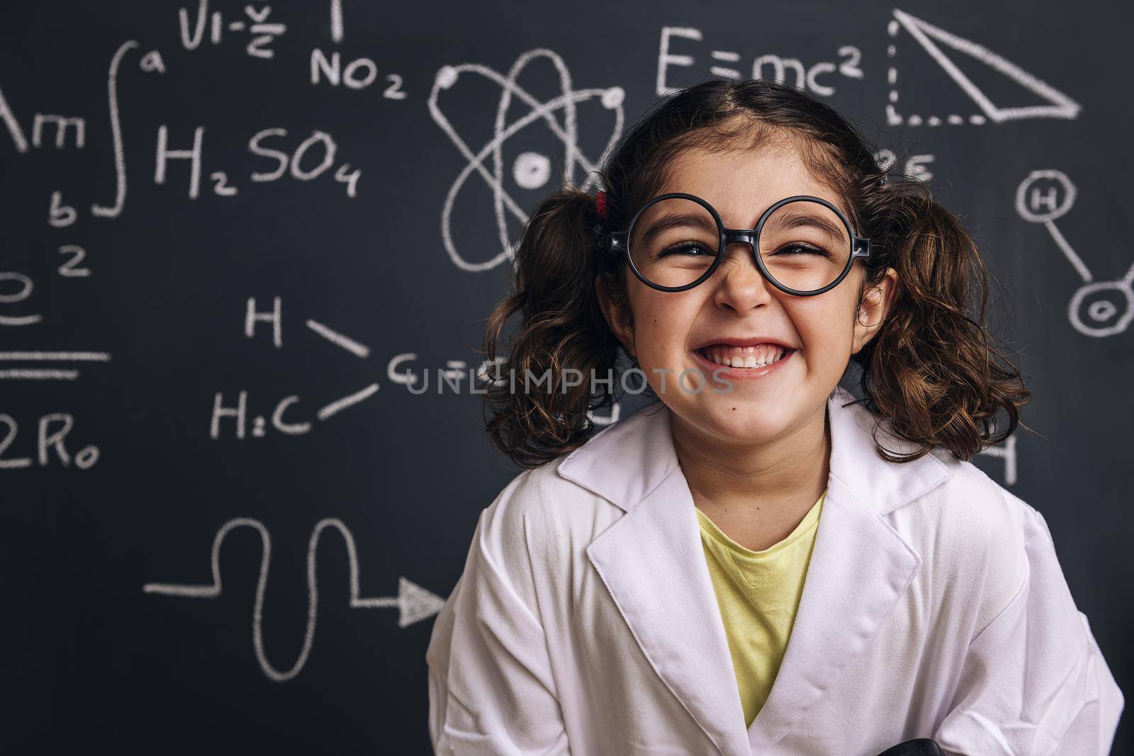 funny little girl science student with glasses in lab coat on school blackboard background with hand drawings science formula pattern, back to school and successful female career concept