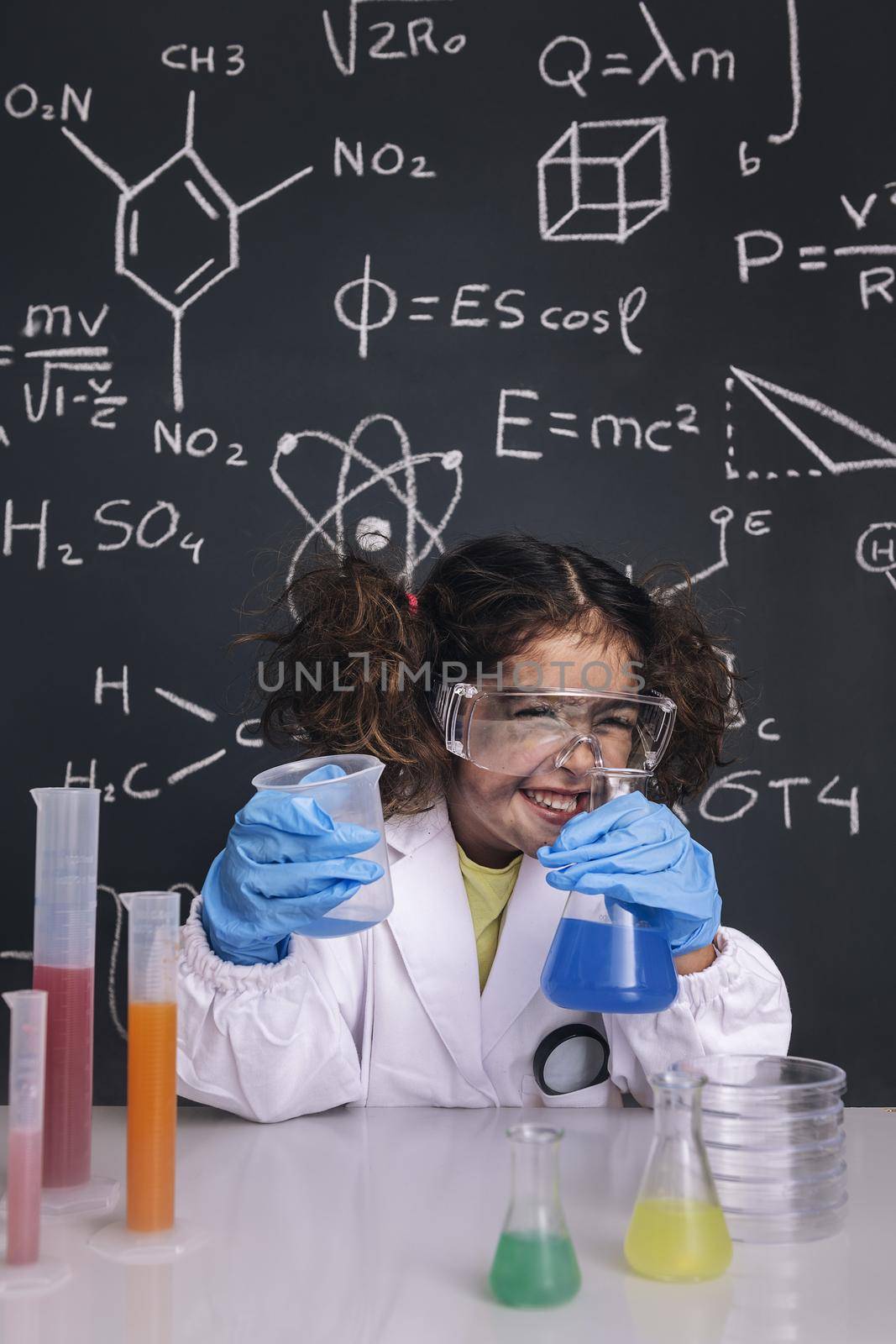 funny scientist girl with gloves and goggles in lab coat laughing, blackboard background with science formulas, explosion in the laboratory, back to school concept