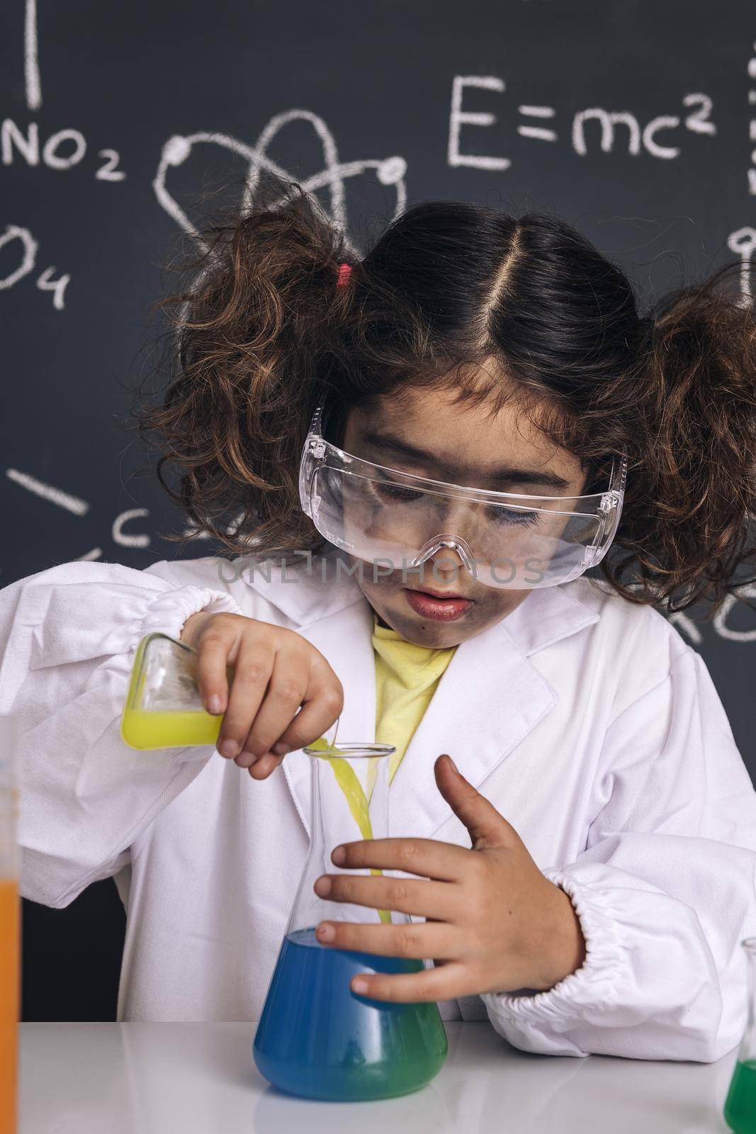 scientist kid with goggles and gloves in lab coat mixing chemical liquids in flasks, blackboard background with science formulas, explosion in the laboratory, back to school concept, vertical photo