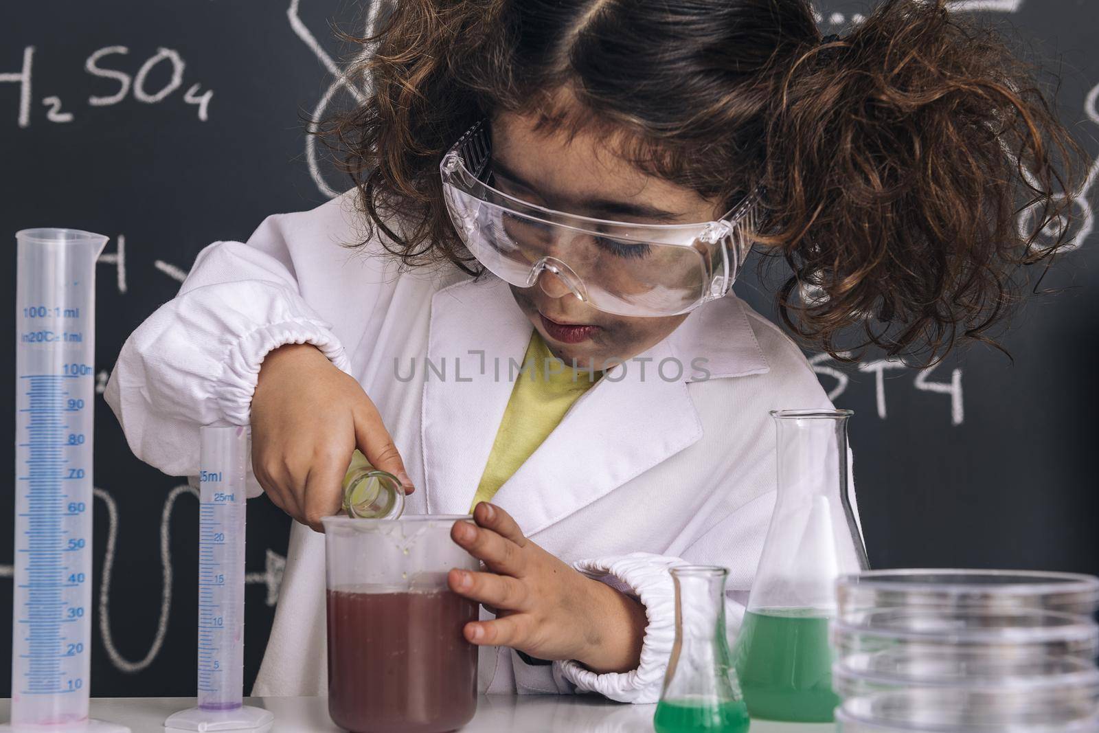 focused scientist child with goggles and gloves in lab coat mixing chemical liquids in flasks, blackboard background with science formulas, explosion in the laboratory, back to school concept